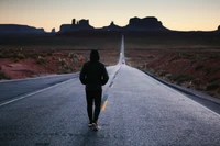 Une silhouette solitaire marchant le long d'une route sinueuse dans Monument Valley à l'aube, encadrée par des formations rocheuses dramatiques et un vaste horizon.