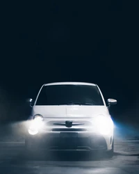 A sleek white compact car illuminated by bright headlights, set against a dark backdrop with subtle fog.