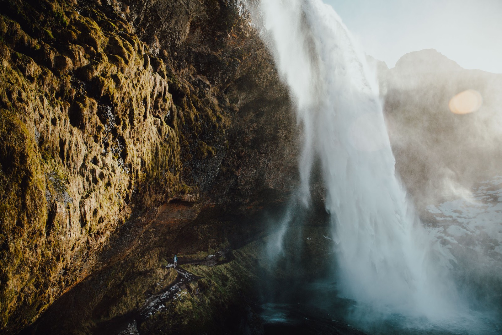 Un homme se tenant devant une cascade avec le soleil qui brille dessus (la cascade, seljalandsfoss, plan deau, nature, eau)