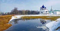 A serene winter landscape featuring a stunning white church with blue domes reflected in a partially frozen pond, surrounded by patches of snow and grass.