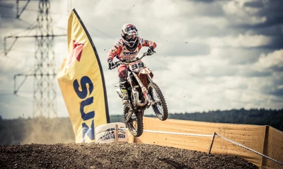 Freestyle Motocross Rider Performing a Jump Over Soil Track with Sponsorship Banner in Background