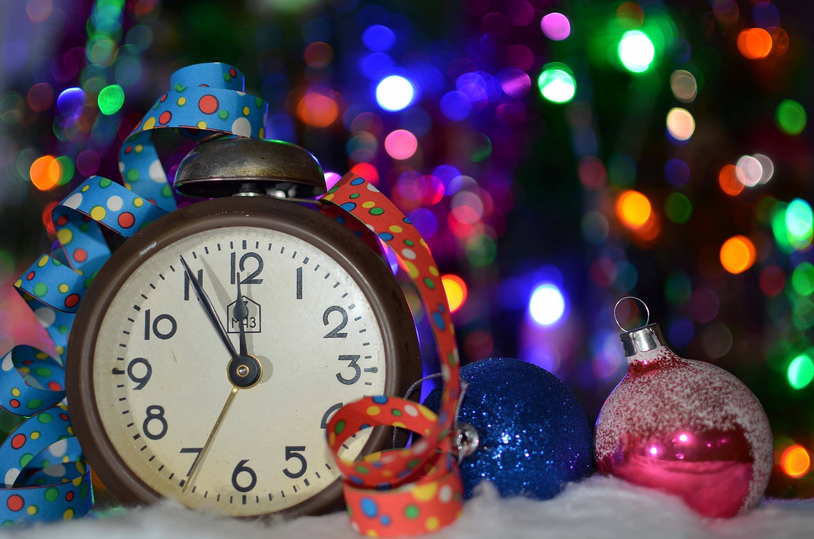 There is a clock and ornaments on a table with a christmas tree in the background (christmas lights, christmas ornament, christmas day, new year, christmas decoration)