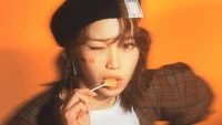 Chaewon playfully winks while holding a lollipop, showcasing a vibrant orange background and a stylish outfit with a beret, capturing a youthful and fun spirit.