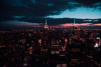 Ein atemberaubender Luftblick auf die Skyline von New York bei Nacht, der beleuchtete Wolkenkratzer zeigt, einschließlich des ikonischen Empire State Buildings, unter einem dramatischen, bewölkten Himmel.