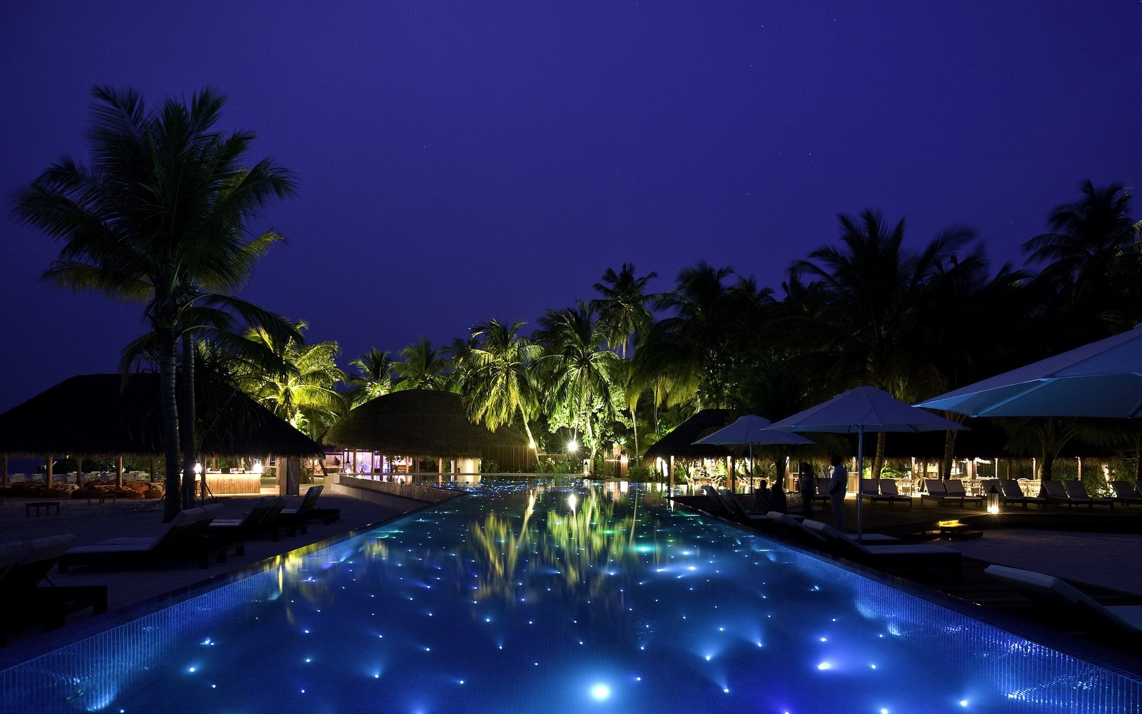 Vista noturna de uma piscina com espreguiçadeiras e guarda-sóis. (piscina, estância, iluminação, villa, noite)