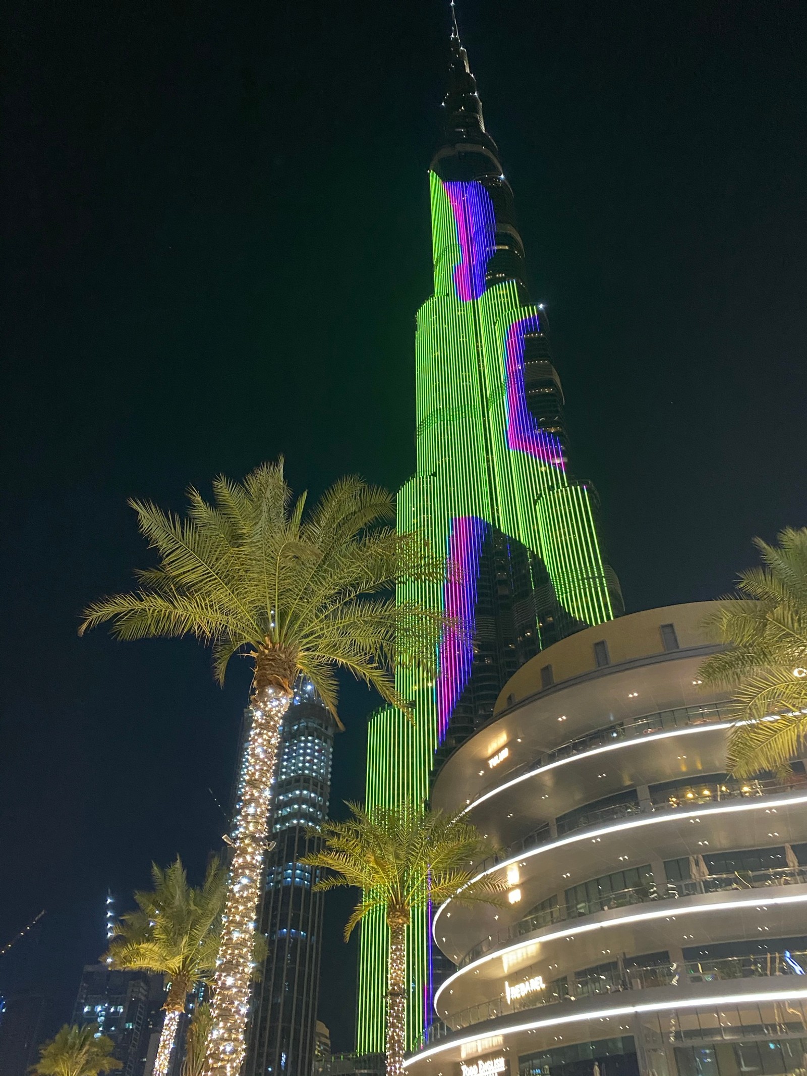 Girafas iluminadas em verde e roxo à noite (torre, palmeiras, azul majorelle, fachada, noite)