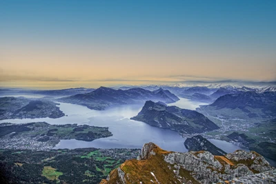 Luftaufnahme des Vierwaldstättersees, umgeben von den Schweizer Alpen, mit schneebedeckten Gipfeln und einer ruhigen Landschaft bei Dämmerung.