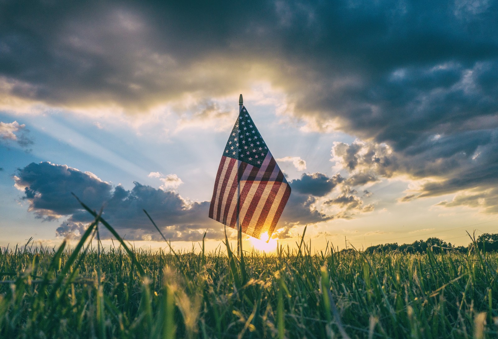 Uma bandeira americana no meio de um campo com o sol brilhando através das nuvens (nuvem, grama, manhã, família das gramíneas, luz solar)