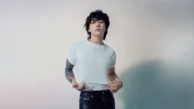 Jungkook dans un haut blanc élégant, mettant en valeur son bras tatoué contre un fond doux et flou.