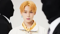 Sunoo from ENHYPEN, featuring a striking orange hairstyle, poses confidently in a white jacket with shadowy figures in the background, symbolizing the concept of "Manifesto: Day 1.