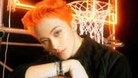 Mark Lee von NCT U mit auffälligen orangefarbenen Haaren, posierend vor einem lebhaften Hintergrund mit Basketball-Elementen und Neonlichtern.