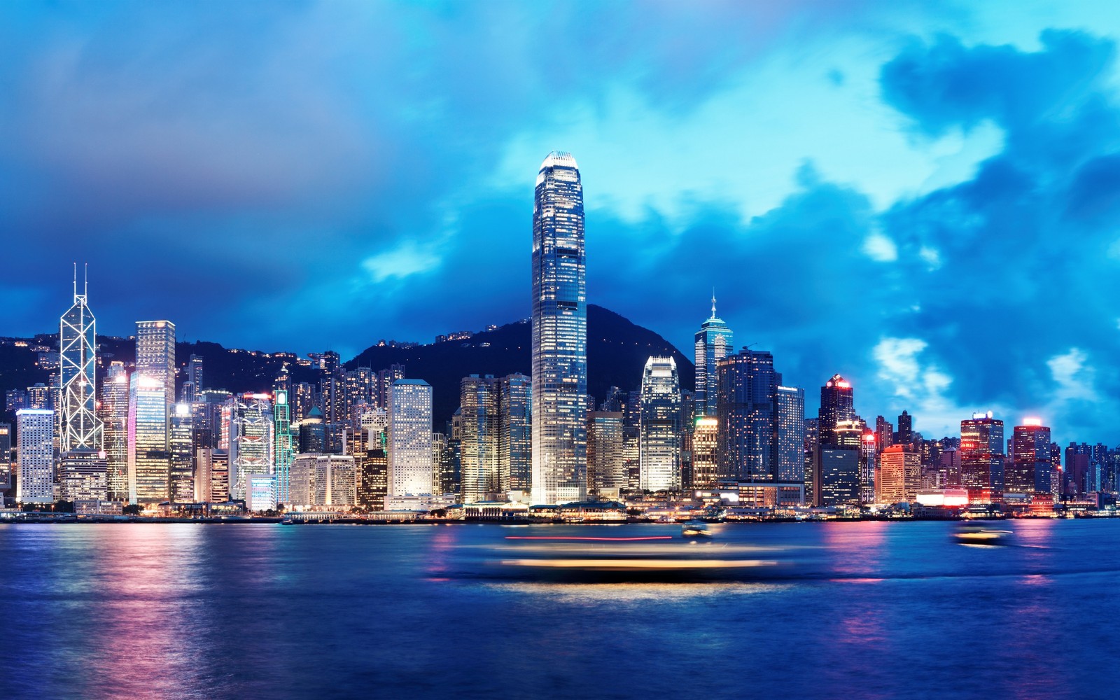 Uma vista borrada do horizonte de uma cidade à noite com um barco em primeiro plano (victoria harbour, cidade, paisagem urbana, linha do horizonte, arranha céus)