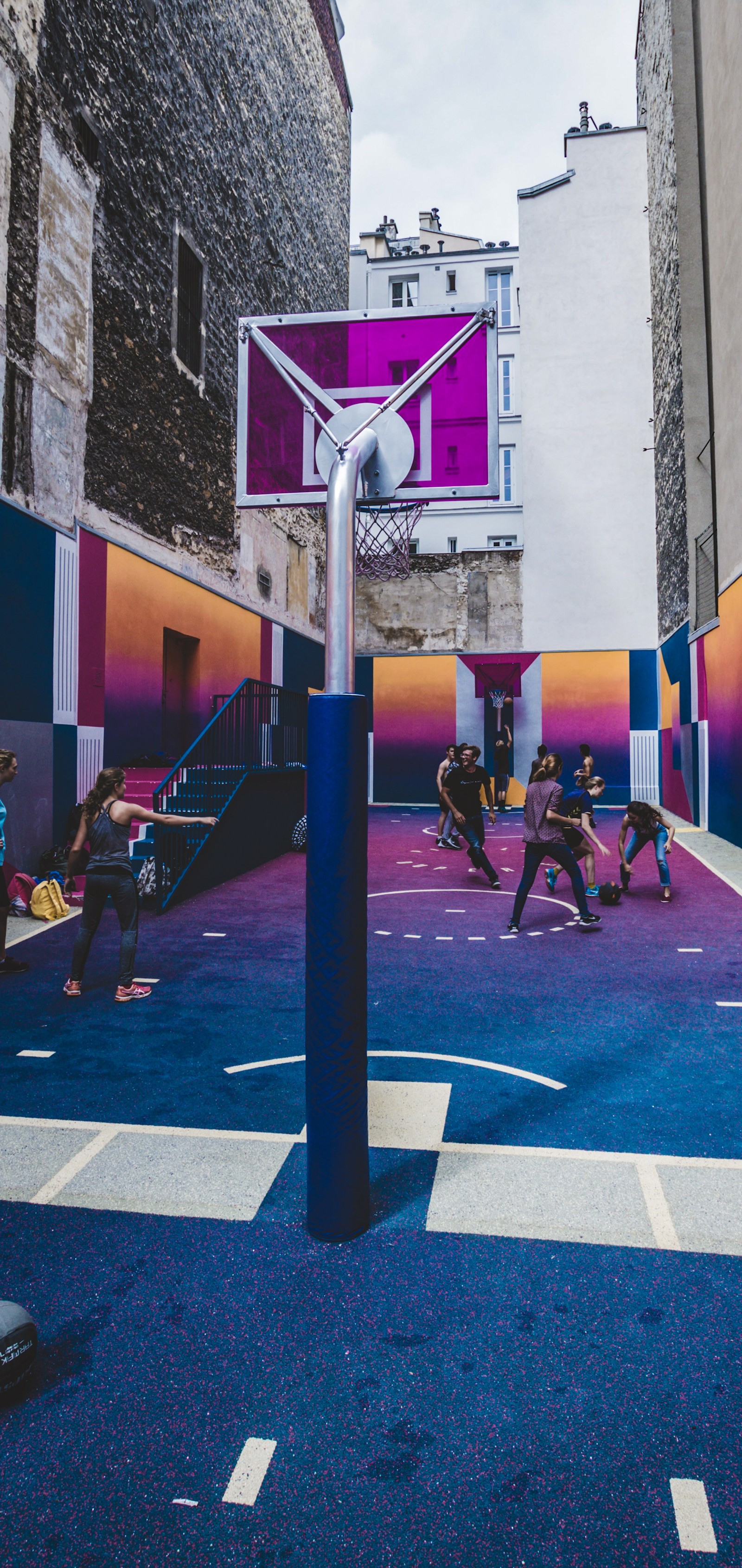 Varias personas están jugando baloncesto en una cancha colorida en una ciudad. (calle, baloncesto, aro de baloncesto, edificio, púrpura)