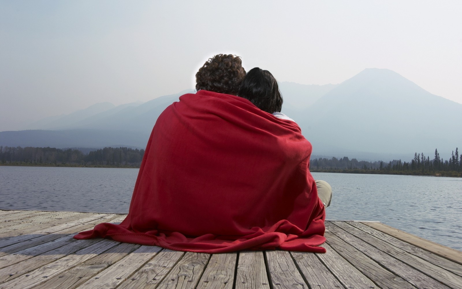 Il y a deux personnes assises sur un quai enveloppées dans une couverture (romance, câlin, couple, rouge, assis)