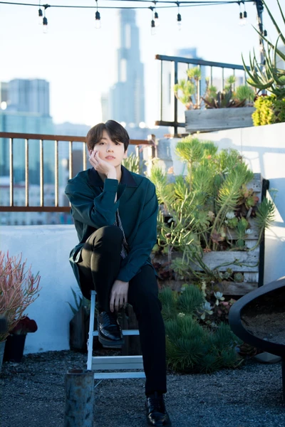 Jungkook de BTS assis pensivement dans un jardin urbain, avec des gratte-ciel en arrière-plan.