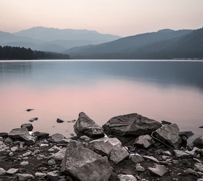 Lago tranquilo al anochecer con primer plano rocoso y siluetas de montañas