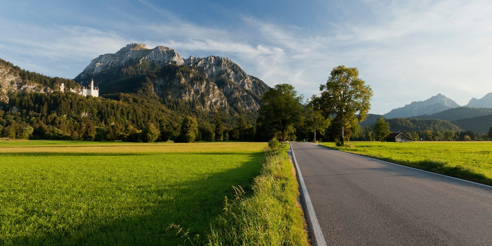 Uma vista de uma estrada no meio de um campo com montanhas ao fundo (castelo de neuschwanstein, castelo, natureza, formas montanhosas, estrada)