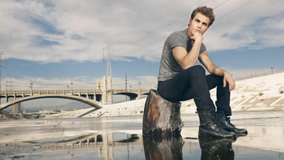 Stefan Salvatore posando pensativo perto da água, cercado por um cenário urbano.