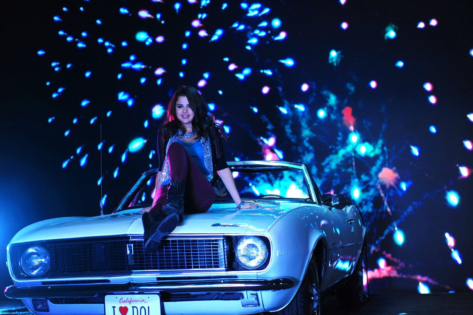 Araffe woman sitting on the hood of a car with fireworks in the background (selena gomez, blue, car, light, headlamp)