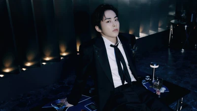 Xiumin d'EXO dans un costume élégant, assis avec une expression sophistiquée dans un cadre élégant avec des boissons et un éclairage d'ambiance.