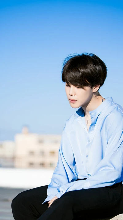 Jimin do BTS em uma pose contemplativa contra um céu azul claro.