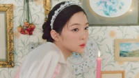 Irene of Red Velvet poses elegantly in a whimsical setting adorned with vintage decor, featuring delicate floral patterns, framed artwork, and a soft candlelight glow.