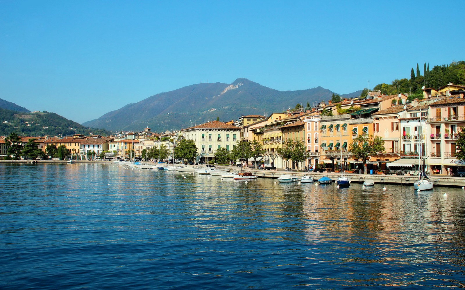 Barcos estão atracados na água em frente a uma fileira de edifícios (lago de garda, lago, cidade, mar, costa)