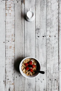 Uma tigela de cereal de café da manhã coberta com morangos e mirtilos frescos, acompanhada por um pequeno jarro de leite, contra um fundo de madeira rústica.