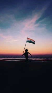 Silhouette of a child waving the Indian tricolor flag against a vibrant sunset at the beach.