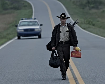 Um xerife caminhando por uma estrada deserta, carregando uma bolsa de duffel e um galão de gasolina, com um carro de polícia ao fundo.