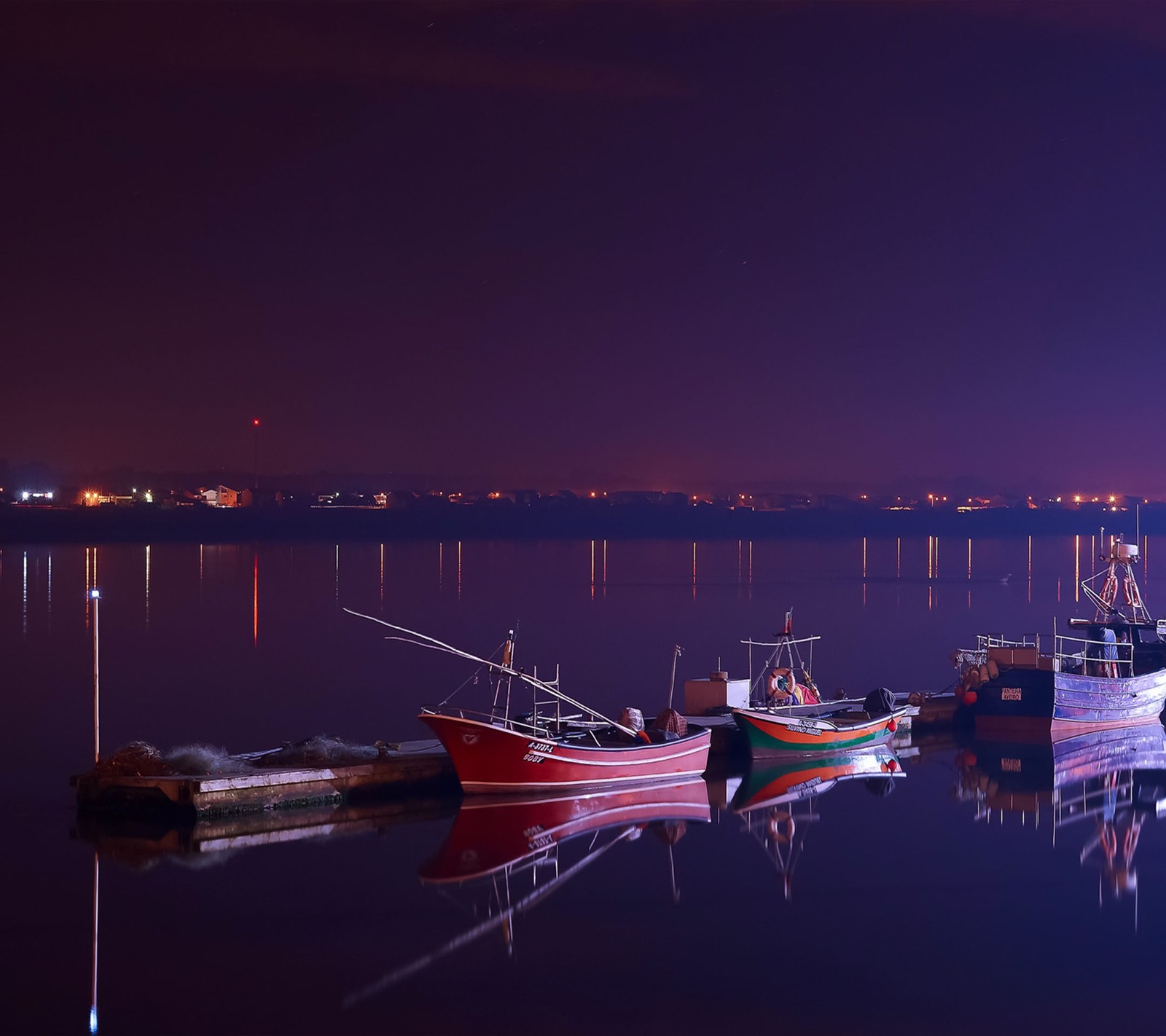 Several boats are docked at a dock at night with a city in the background (boats, two)