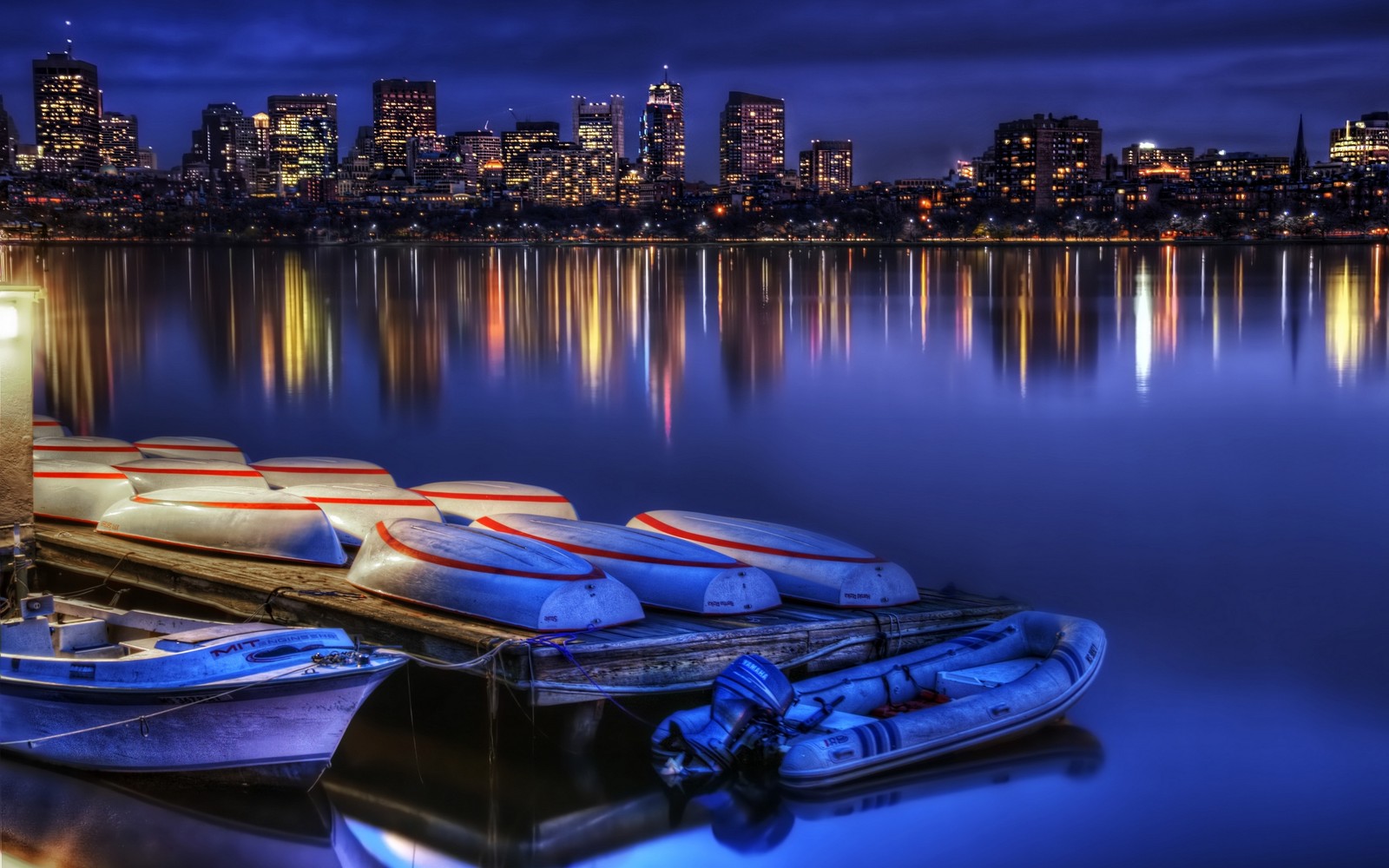 Los barcos están alineados en un muelle frente a un horizonte urbano (transporte acuático, reflexión, ciudad, noche, agua)