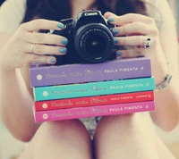 A girl holds a camera while sitting with colorful books stacked in front of her.