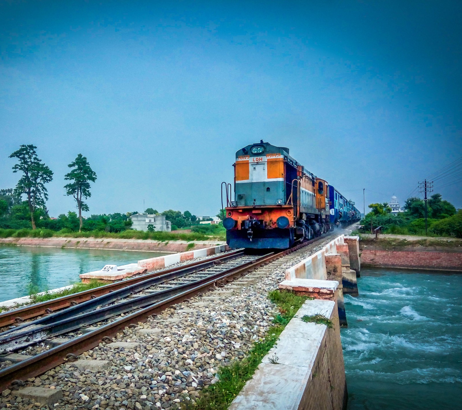 Un tren que avanza por las vías junto al agua (azul, flores, verde, railtracks, ferrocarril)