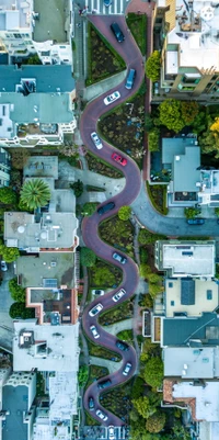 Aerial view of Lombard Street's iconic zigzag pathway, surrounded by residential areas and lush greenery, with glimpses of urban design and nature.