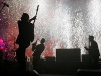 Desempenho dinâmico em um concerto de rock progressivo, apresentando músicos silhuetados contra um fundo de fogos de artifício vibrantes.