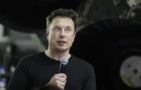 Elon Musk speaking with a microphone in front of a spacecraft.