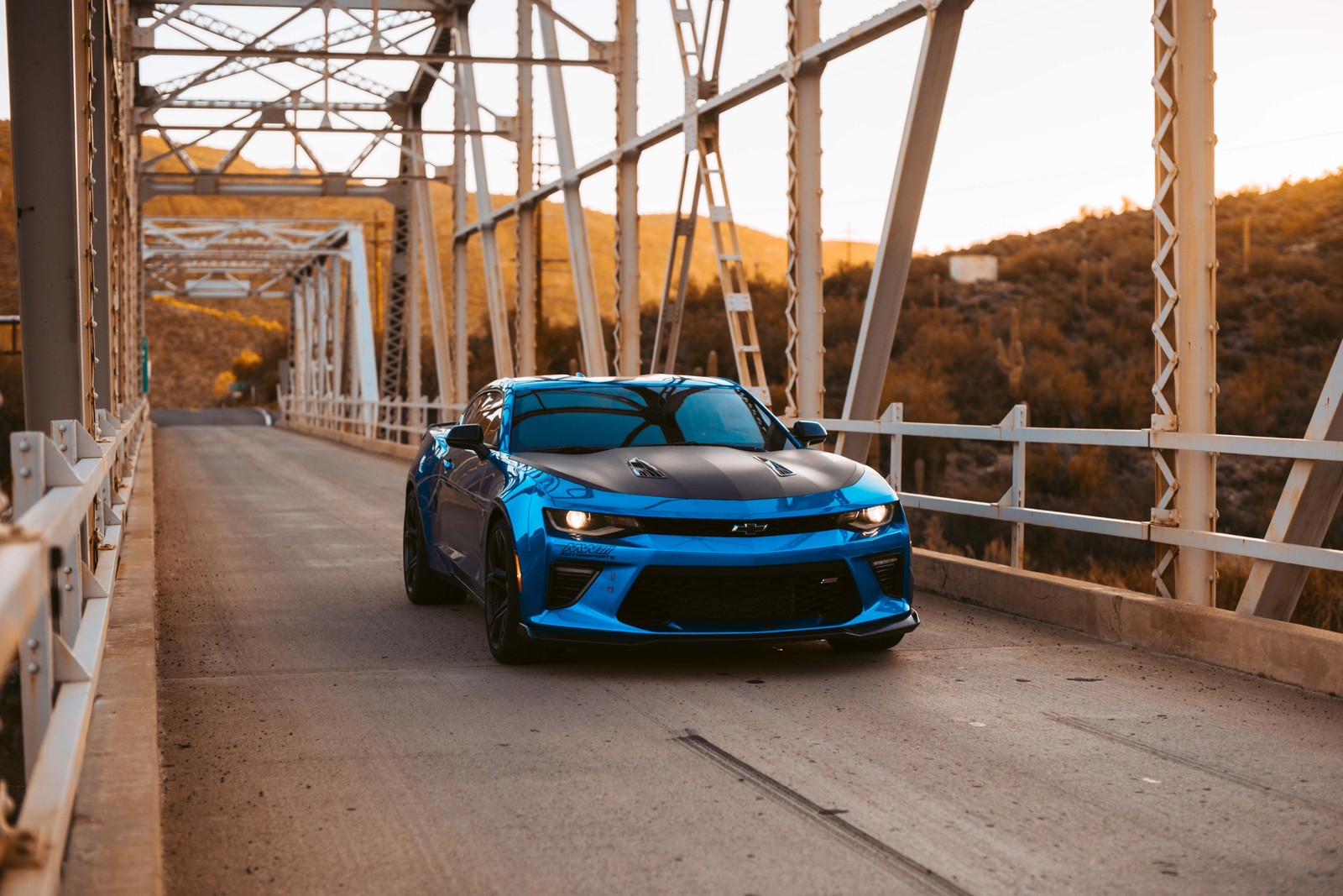 A blue car driving across a bridge with mountains in the background (car, chevrolet, muscle car, bumper, automotive exterior)