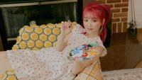 Seunghee do Oh My Girl, segurando brincando um pincel e uma paleta, vestida com um vestido branco floral, com um cabelo rosa vibrante, contra um fundo aconchegante e bem iluminado.