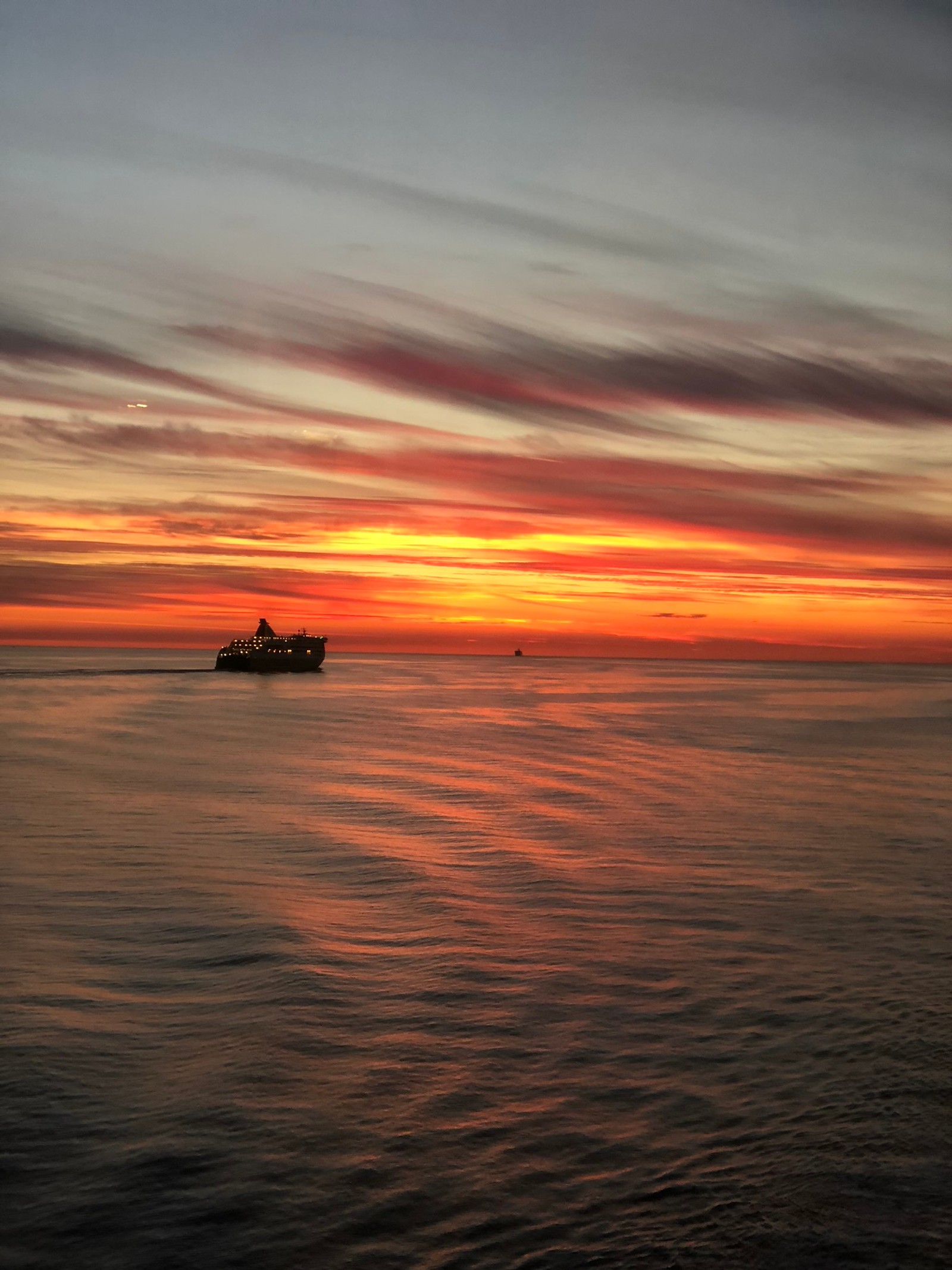 Uma vista aérea de um barco no oceano ao pôr do sol com um navio ao longe (horizonte, água, por do sol, corpo de água, sol)