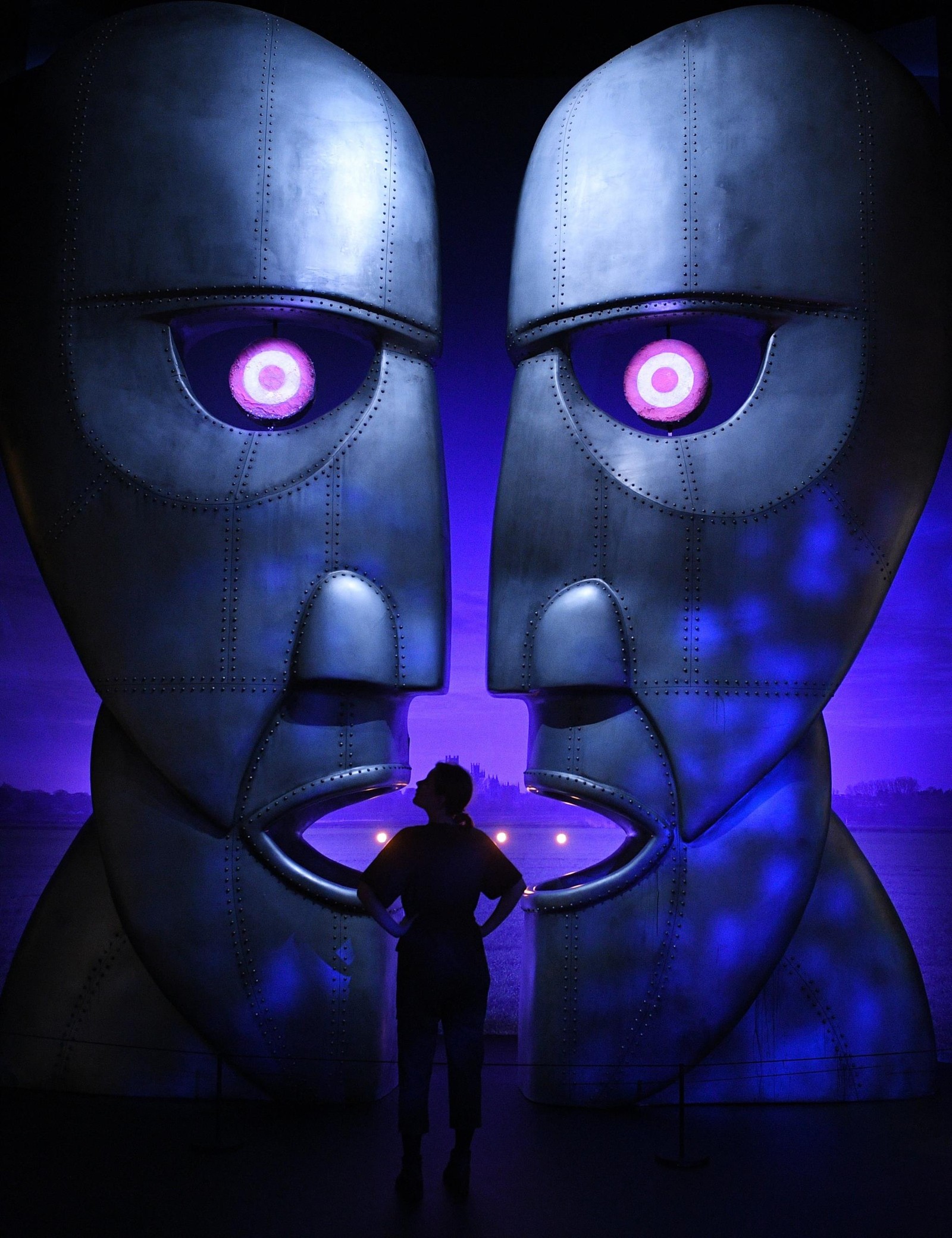 Un homme se tient devant une grande sculpture de deux visages (pink floyd, the division bell, violet, obscurité, symétrie)