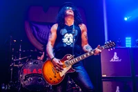 Slash tritt auf der Bühne mit einer E-Gitarre während eines Rockkonzerts auf, mit lebhaften Lichtern und einem Hintergrund mit Bandbildern.