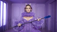 Katy Perry in einem auffälligen lila Outfit mit einem dramatischen Kragen, hält einen blauen Stab vor einem passenden lila Hintergrund.