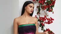 Camila Mendes in a vibrant strapless dress, posed against a backdrop of blooming red flowers.