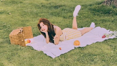 Seulgi relaxando em uma toalha de piquenique cercada por frutas em um campo verdejante.