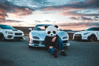 Carros esportivos de médio porte sob um céu colorido com um entusiasta mascarado de panda