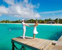 Una pareja romántica tomándose de las manos en un muelle de madera, rodeada de aguas turquesas y un paisaje tropical soleado.