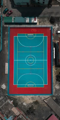 Vista aérea de uma quadra de futsal vibrante com um design simétrico em azul e vermelho, cercada por uma mistura de estruturas urbanas e espaços verdes.