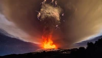 Espectáculo eruptivo del Monte Etna, con un domo de lava ardiente y una nube de ceniza que se eleva iluminada por relámpagos, capturando la intensidad de una explosión volcánica en medio de un cielo dramático.