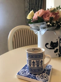 Taza de café de cerámica con diseño griego al lado de un arreglo floral en una mesa.